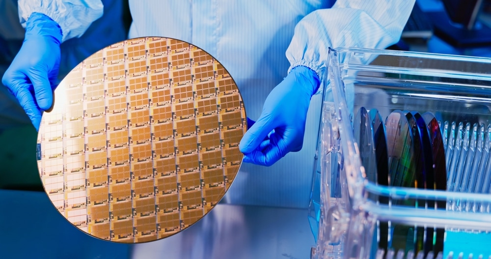 Técnico de jaleco e luvas azuis segura um wafer de silício dourado com chips, ao lado de um estojo com outros wafers em uma fábrica de semicondutores