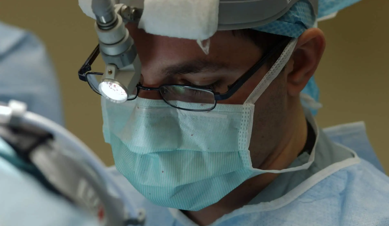 Profissional de saúde de máscara cirúrgica, óculos e lanterna de cabeça, concentrado em um procedimento em sala de cirurgia