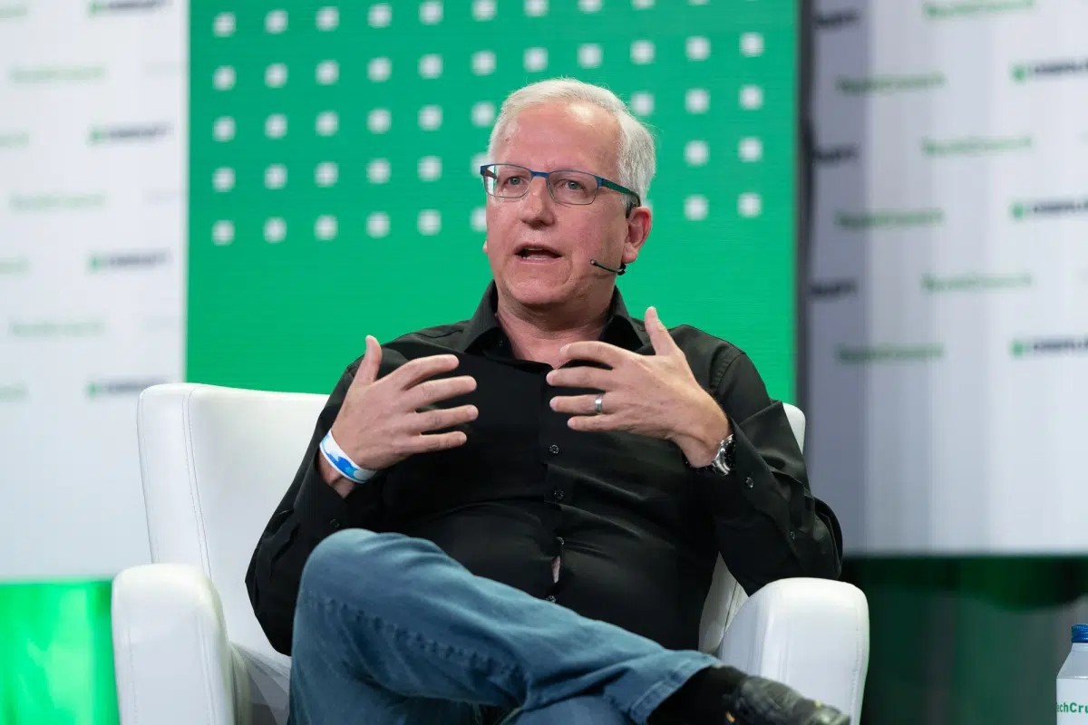 Homem de óculos e camisa preta, sentado em poltrona branca, gesticulando durante painel em evento de tecnologia com fundo verde