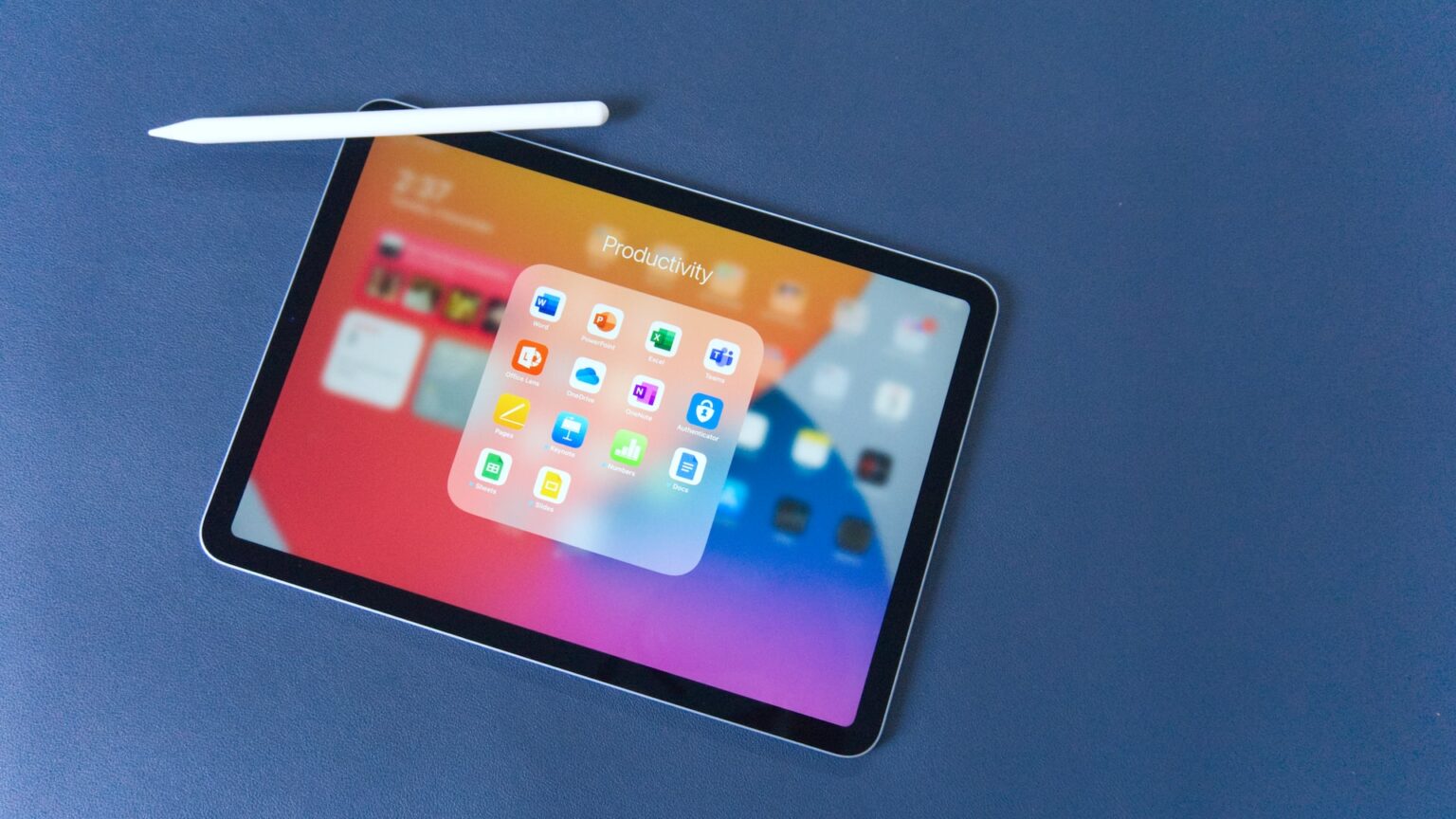 Tablet da Apple com caneta stylus branca sobre superfície azul, exibindo pasta "Productivity" com aplicativos de escritório na tela