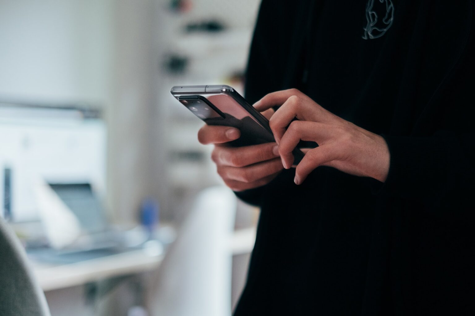 Pessoa de roupa preta usando smartphone com as duas mãos, em ambiente de escritório desfocado ao fundo
