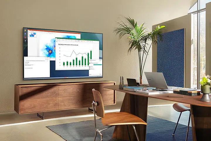 TV Samsung Crystal UHD fixada na parede de um escritório, exibindo planilha com gráfico de barras e linhas, com mesa de madeira, notebook e planta ao lado