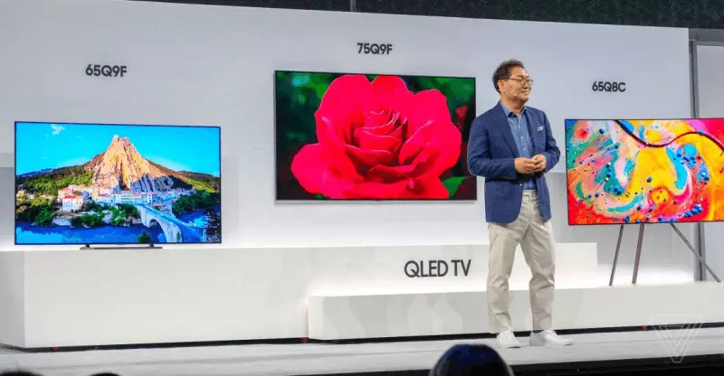 Apresentação de TVs QLED em palco, com três modelos exibidos: 65Q9F, 75Q9F e 65Q8C, e um apresentador de blazer ao lado