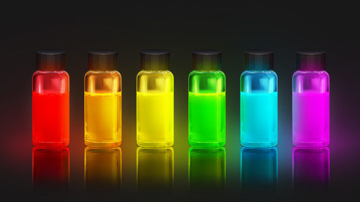 Seis frascos de vidro com líquidos coloridos em vermelho, laranja, amarelo, verde, ciano e magenta, sobre fundo escuro com reflexos