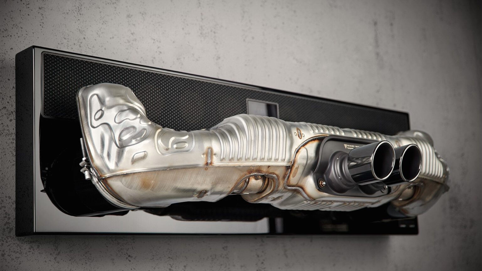 Soundbar em formato de escapamento esportivo de Porsche, com duas ponteiras cromadas, montada na parede sobre base preta de acabamento espelhado