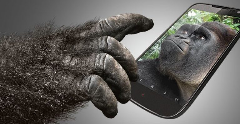 Mão de gorila saindo da tela de um smartphone, com um gorila olhando para fora pela tela, sobre fundo cinza