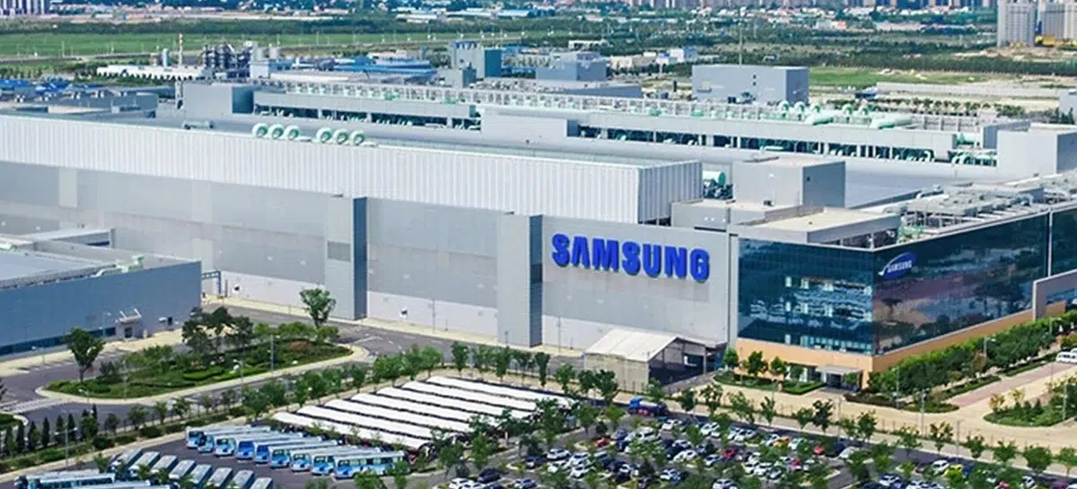 Vista aérea de grande fábrica da Samsung, com prédios cinza, logo azul da marca na fachada e vasto estacionamento com veículos
