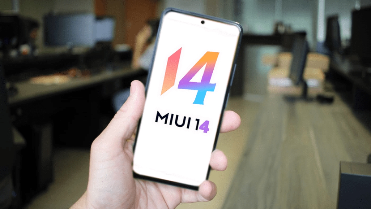 Mão segurando smartphone exibindo o logotipo colorido do MIUI 14 na tela, com escritório desfocado ao fundo