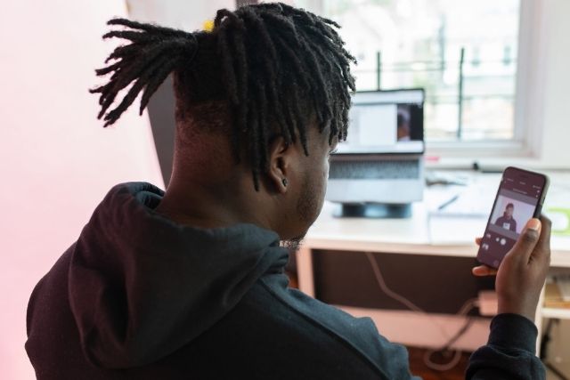 Homem de costas, com dreads, segura celular mostrando uma videochamada enquanto um notebook está aberto ao fundo