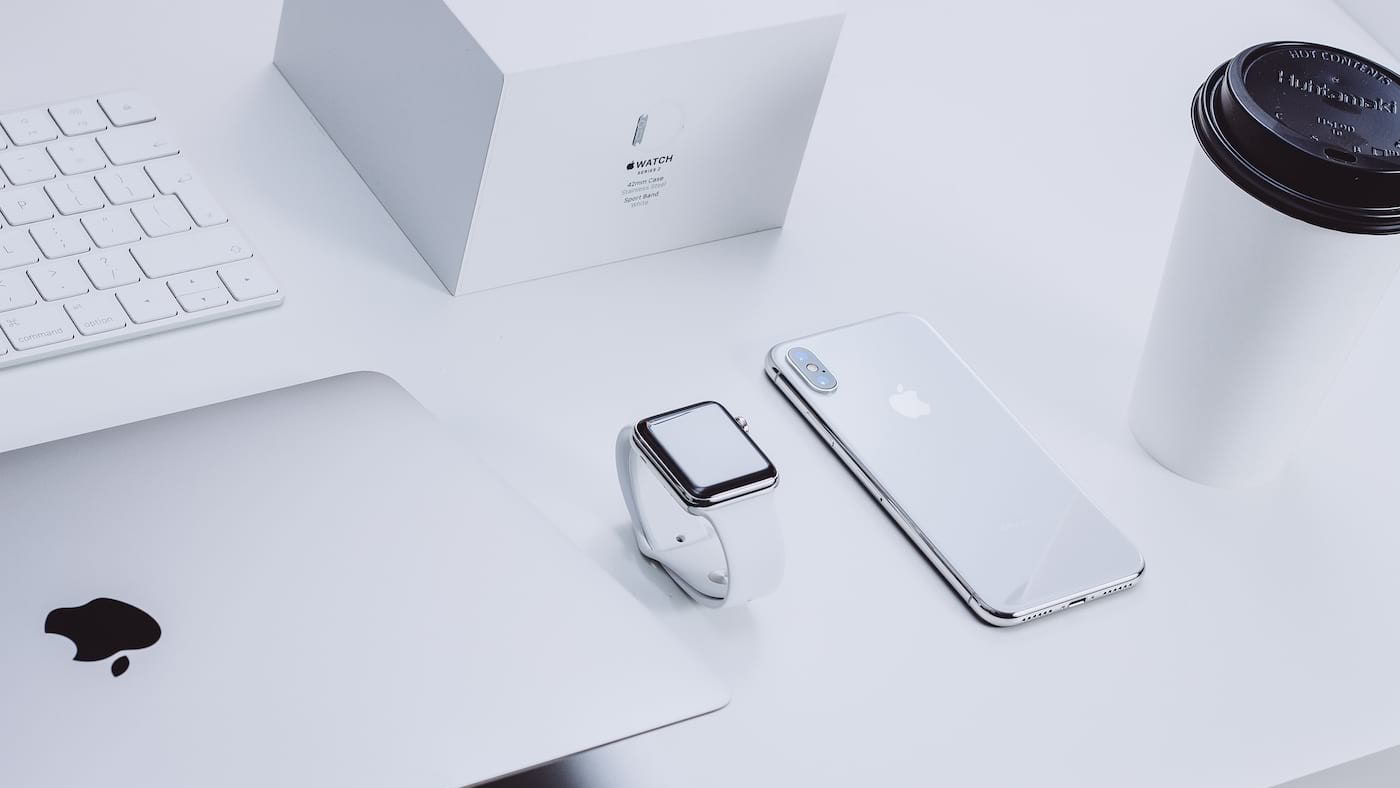 Mesa branca com MacBook, teclado, Apple Watch branco, iPhone prateado e caixa do Apple Watch ao fundo