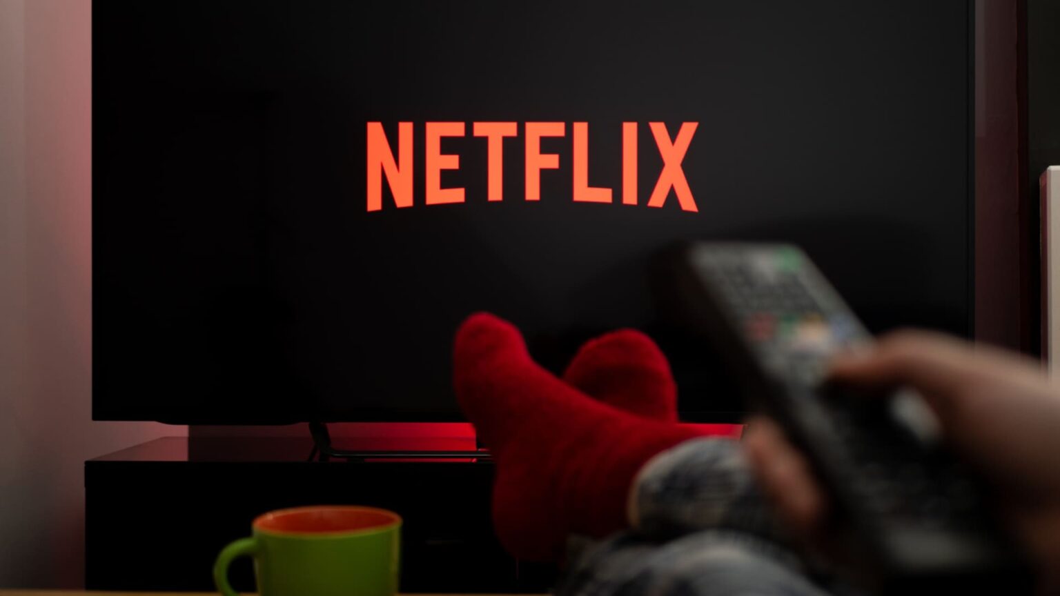 Televisão exibindo o logotipo da Netflix, com pés de meia vermelha apoiados na mesa e uma pessoa segurando um controle remoto em primeiro plano