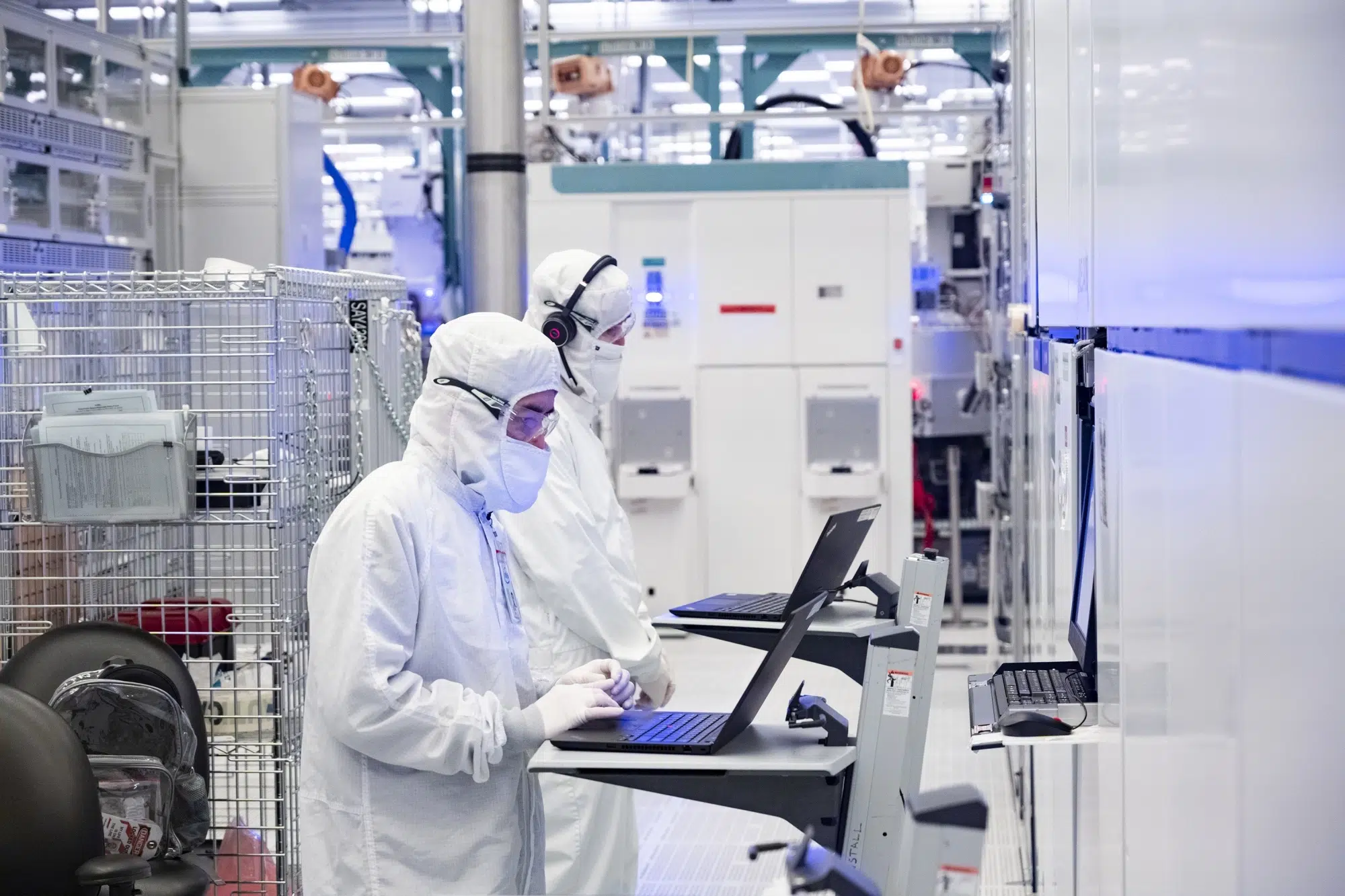 Duas pessoas com roupas brancas de sala limpa, toucas e máscaras, trabalhando em notebooks dentro de uma fábrica de semicondutores com equipamentos automatizad…