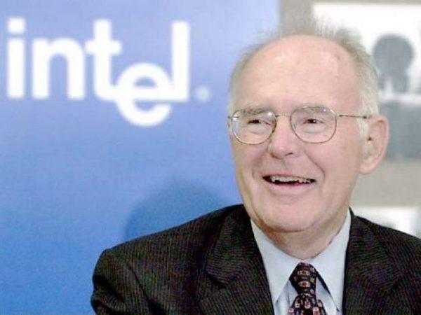 Homem sorrindo de óculos e terno em frente a um painel azul com o logotipo da Intel em branco