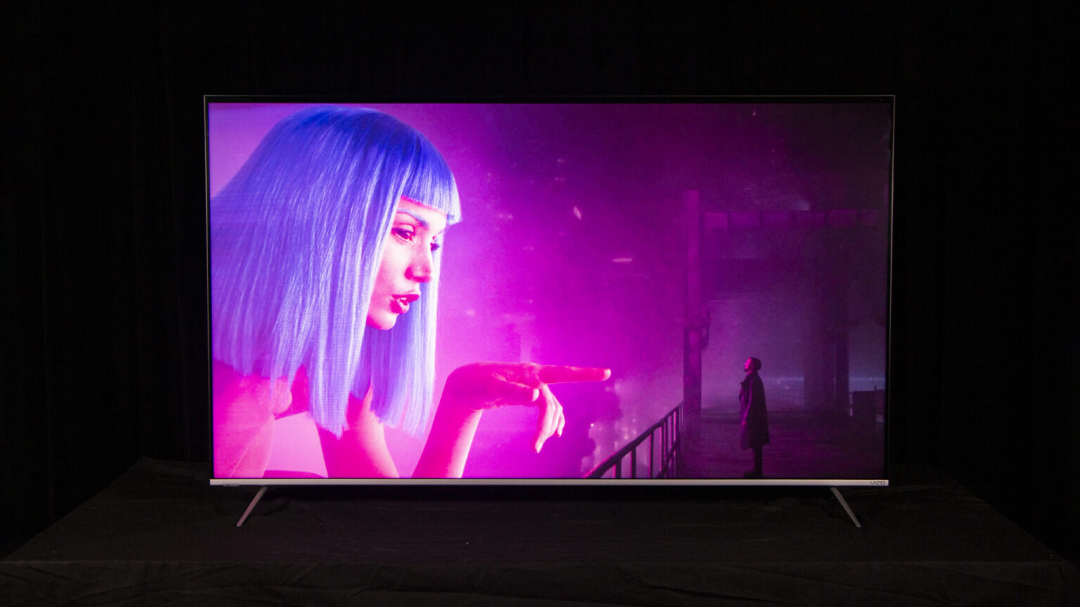 Televisão com tela de alto contraste exibindo cena roxa e rosa com personagem de cabelo azul e silhueta de uma pessoa ao fundo