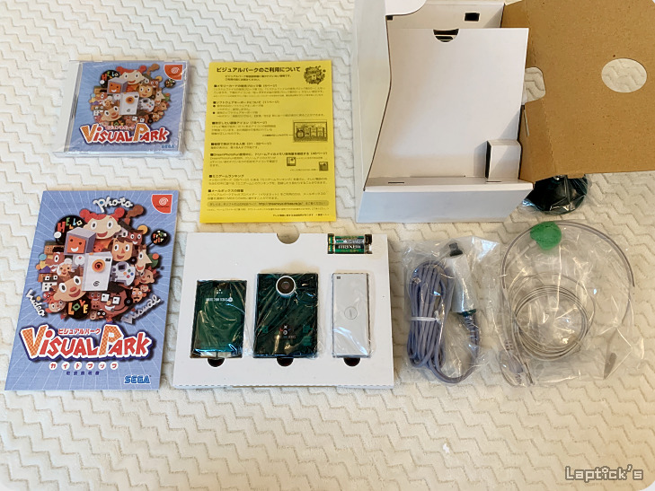 Conteúdo de caixa do Sega Dreamcast Visual Park, com o jogo, guia em japonês, câmera, cabos e acessórios espalhados sobre uma superfície branca