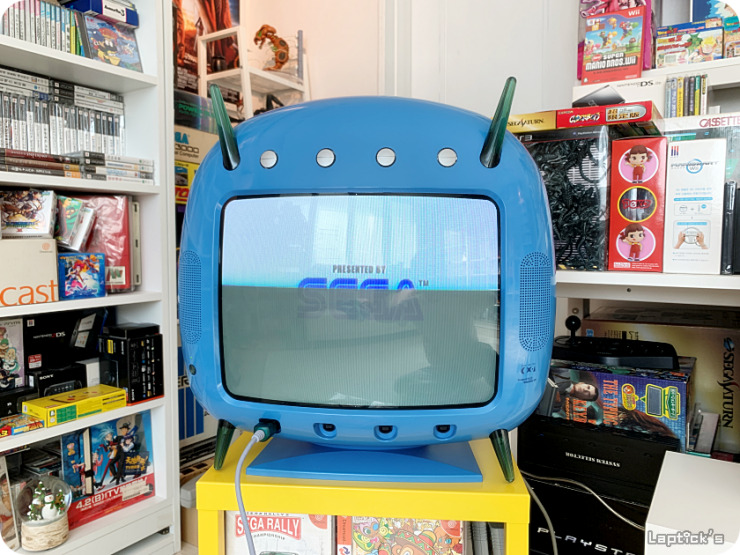 TV azul retrô com tela exibindo a logo "Presented by SEGA", ao lado de estantes com jogos e consoles, incluindo um Sega Saturn e caixas de Dreamcast