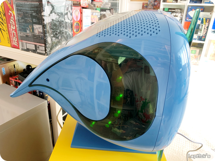 Console Sega Dreamcast embutido em TV azul translúcida de formato arredondado, com LEDs verdes visíveis pela janela lateral, em exibição sobre base amarela