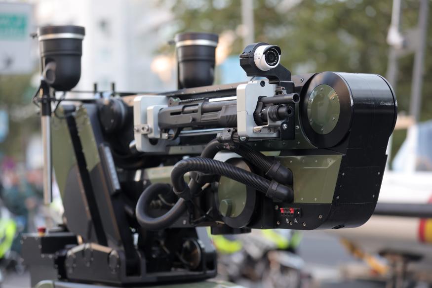 Sistema de armamento remoto montado em veículo militar, com câmeras, sensores e um canhão de tiro automático de cor preta e cinza, em uma rua com pessoas ao fu…