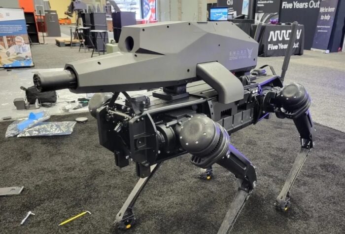 Robô quadrúpede de pernas articuladas com sistema de armas montado no dorso, exibido em estande de feira, da fabricante Ghost Robotics