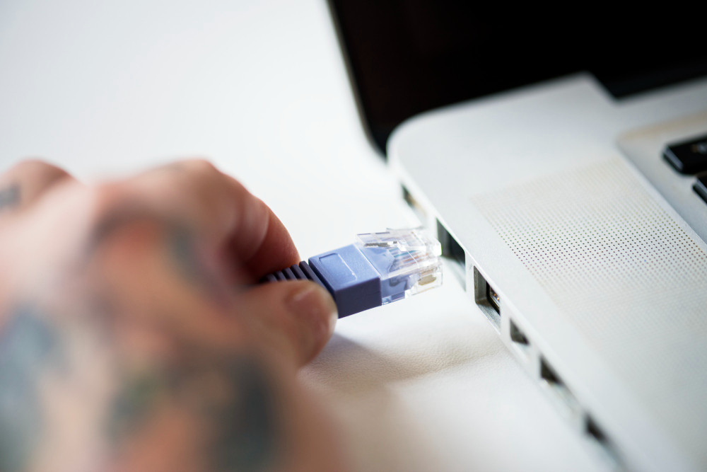Mão conectando um cabo de rede Ethernet azul com conector RJ45 em uma porta de um notebook prateado