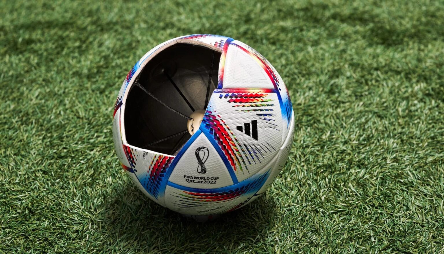 Bola oficial da Copa do Mundo FIFA Qatar 2022 da Adidas, branca com detalhes coloridos, com a seção frontal aberta para mostrar o sensor interno, sobre um gram…