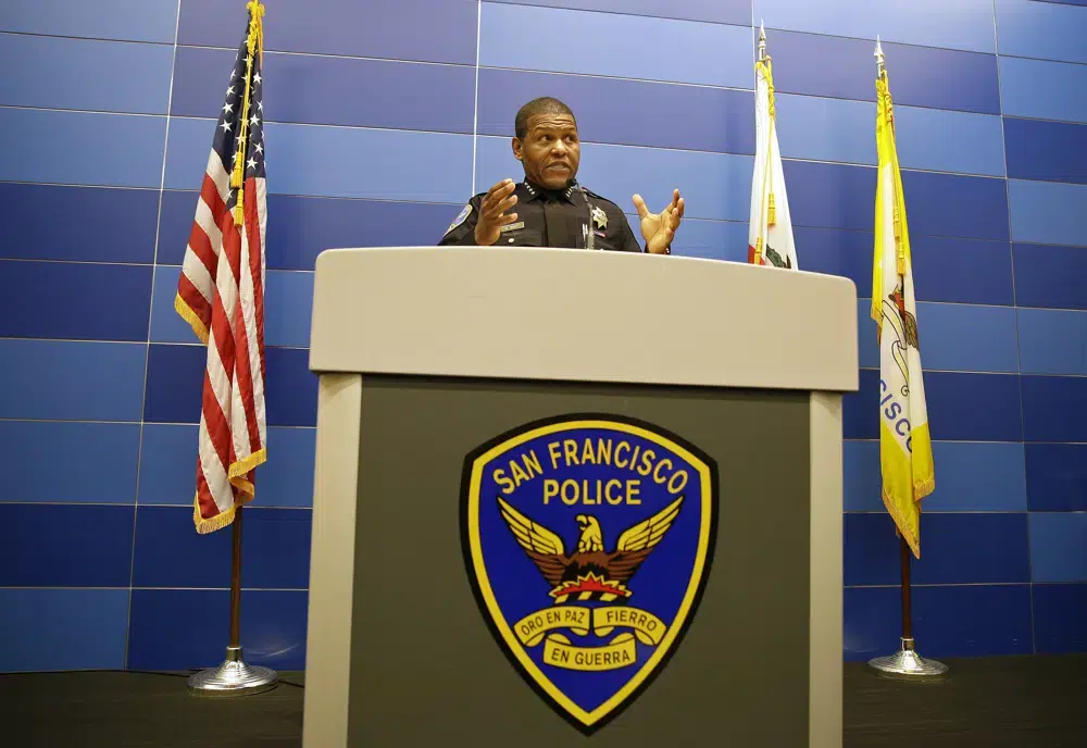 Policial de uniforme em coletiva de imprensa atrás de pódio com brasão da San Francisco Police, ladeado por bandeiras dos EUA e da Califórnia