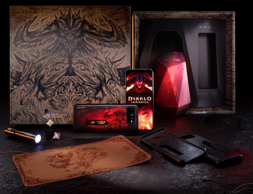 Smartphone ROG Phone 6 Diablo Immortal Edition com tela exibindo o logo do jogo, ao lado da caixa de edição especial, mousepad, lanterna e acessórios decorados…