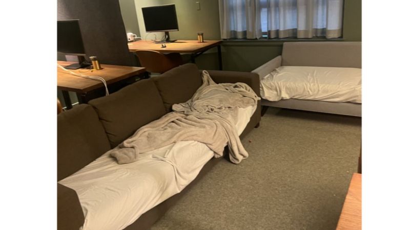 Sofá marrom com lençol e cobertor bagunçados, usado como cama, ao lado de um colchão em estrutura clara, em um escritório com mesa de madeira e monitor ao fundo