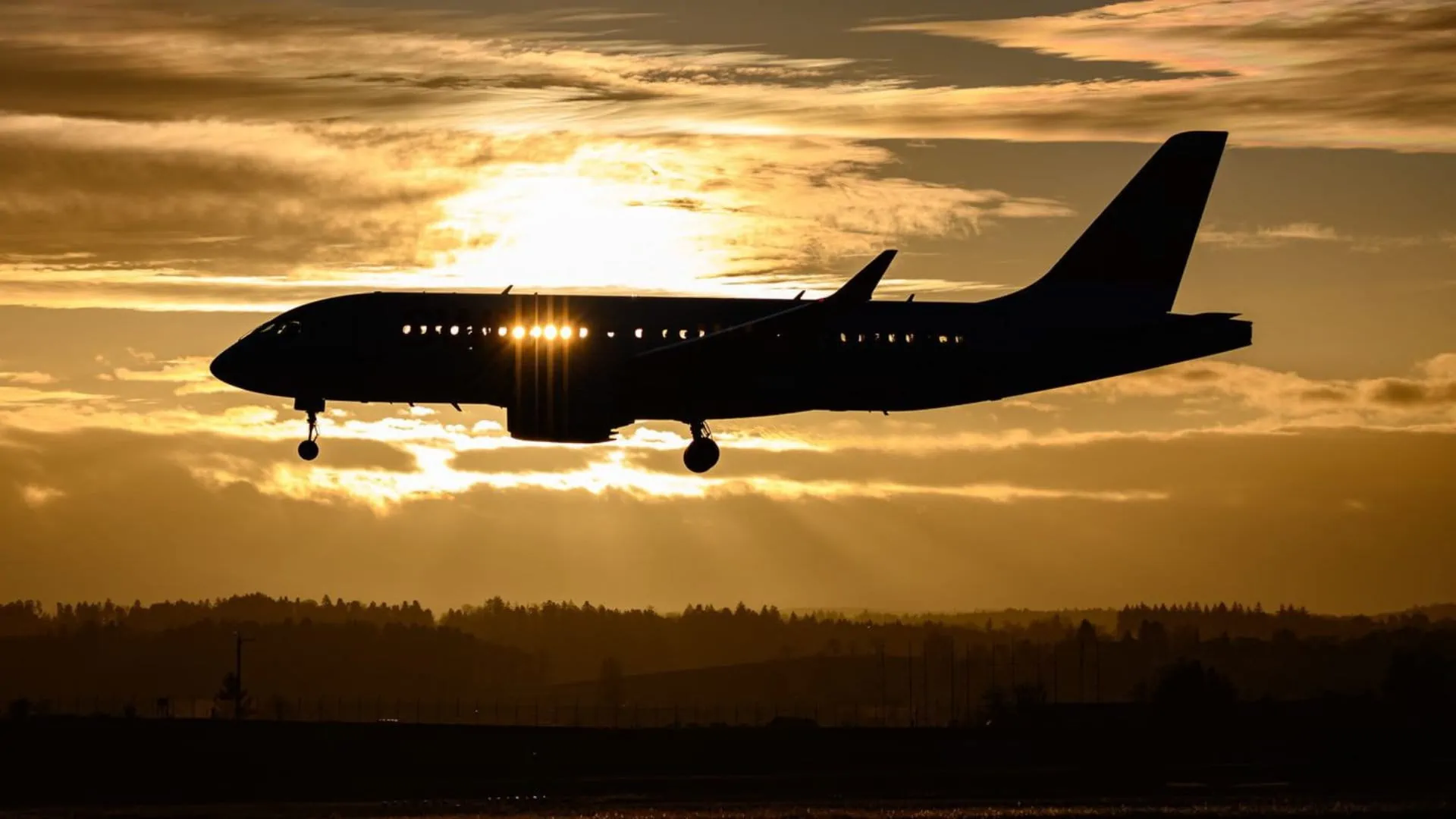 Avião comercial em silhueta pousando ao entardecer, com o trem de pouso estendido e o sol atrás da aeronave, sobre um horizonte de árvores