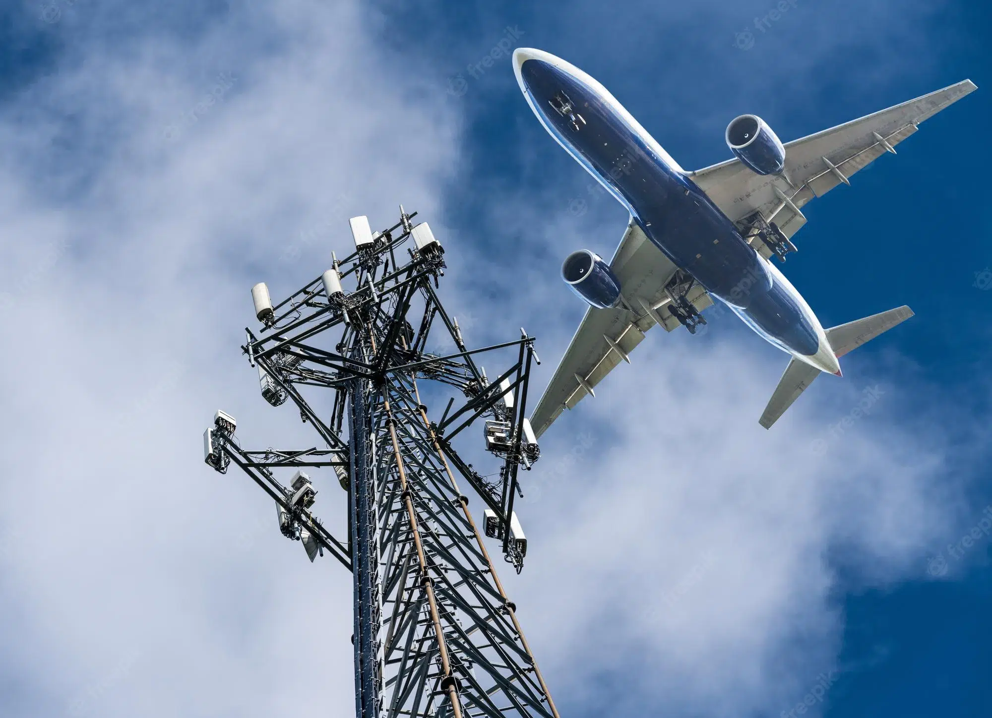 Torre metálica de telefonia celular com antenas 5G em primeiro plano e um avião de passageiros sobrevoando contra o céu azul