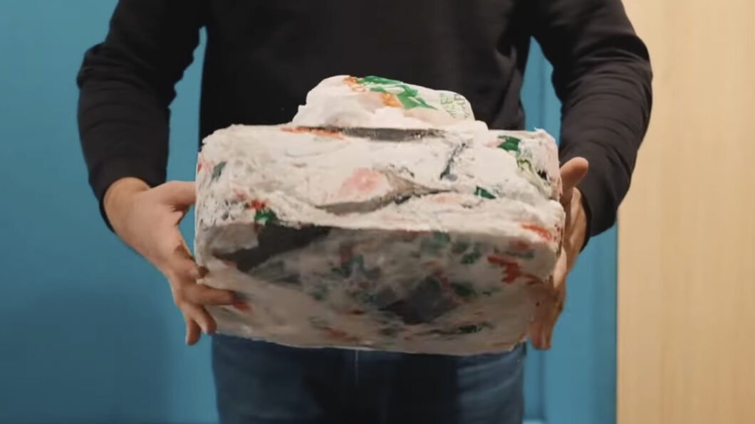 Pessoa segurando um bloco branco e irregular de plástico derretido com restos de embalagens coloridas, em frente a uma parede azul