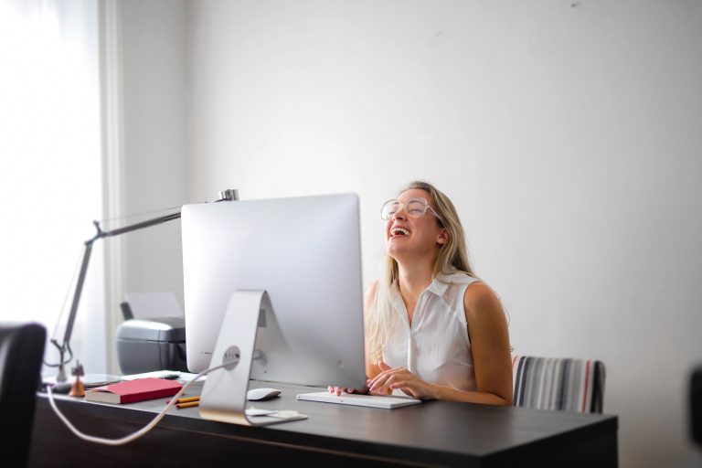 Mulher de óculos sorrindo diante de um computador All in One prateado sobre mesa de madeira escura, em escritório claro