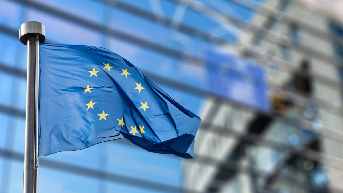 Bandeira da União Europeia tremulando em mastro de metal, com prédio de vidro azul desfocado ao fundo