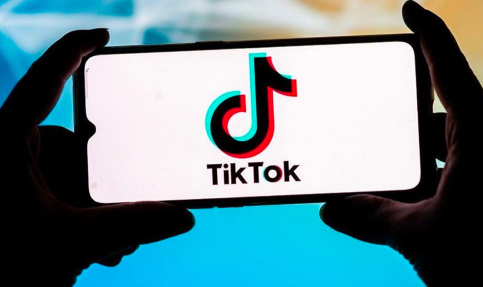 Mãos segurando um smartphone na horizontal, com o logotipo do TikTok exibido na tela branca sobre fundo azul desfocado