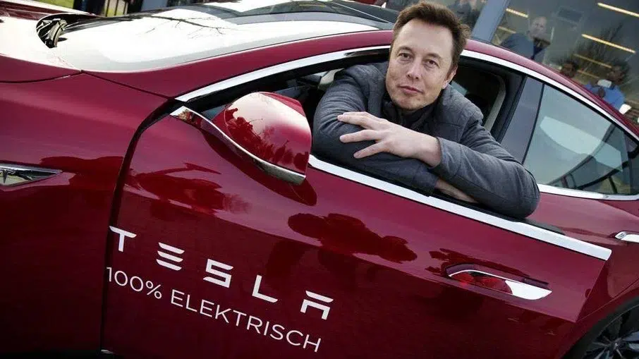 Homem de camisa cinza apoiado na janela de um carro vermelho Tesla com o texto "100% Elektrisch" na porta
