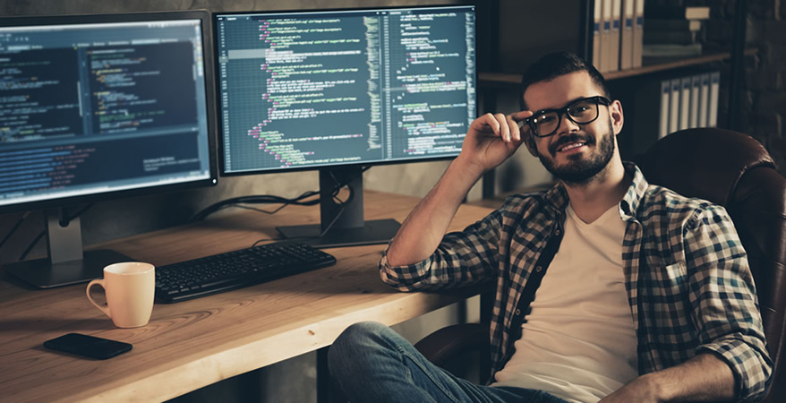 Homem sorridente de óculos sentado em frente a dois monitores exibindo código-fonte, com uma caneca e um teclado sobre a mesa de madeira