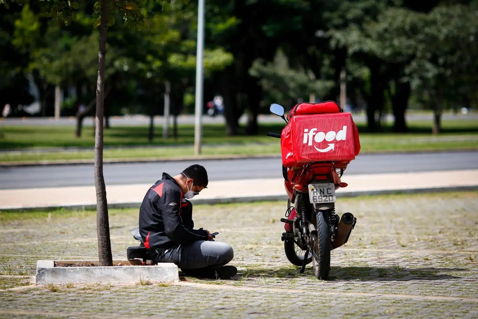 Entregador de moto com baú vermelho da iFood sentado no chão de uma praça, mexendo no celular ao lado da moto