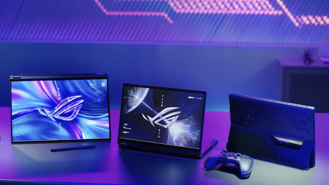 Três notebooks Asus ROG com telas exibindo o logo ROG, lado a lado sobre uma mesa com iluminação roxa e azul, ao lado de um controle de videogame