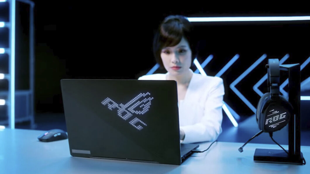 Notebook gamer Asus ROG preto com logo ROG em LEDs na tampa, ao lado de headset ROG e mouse, com uma mulher digitando em cenário azul