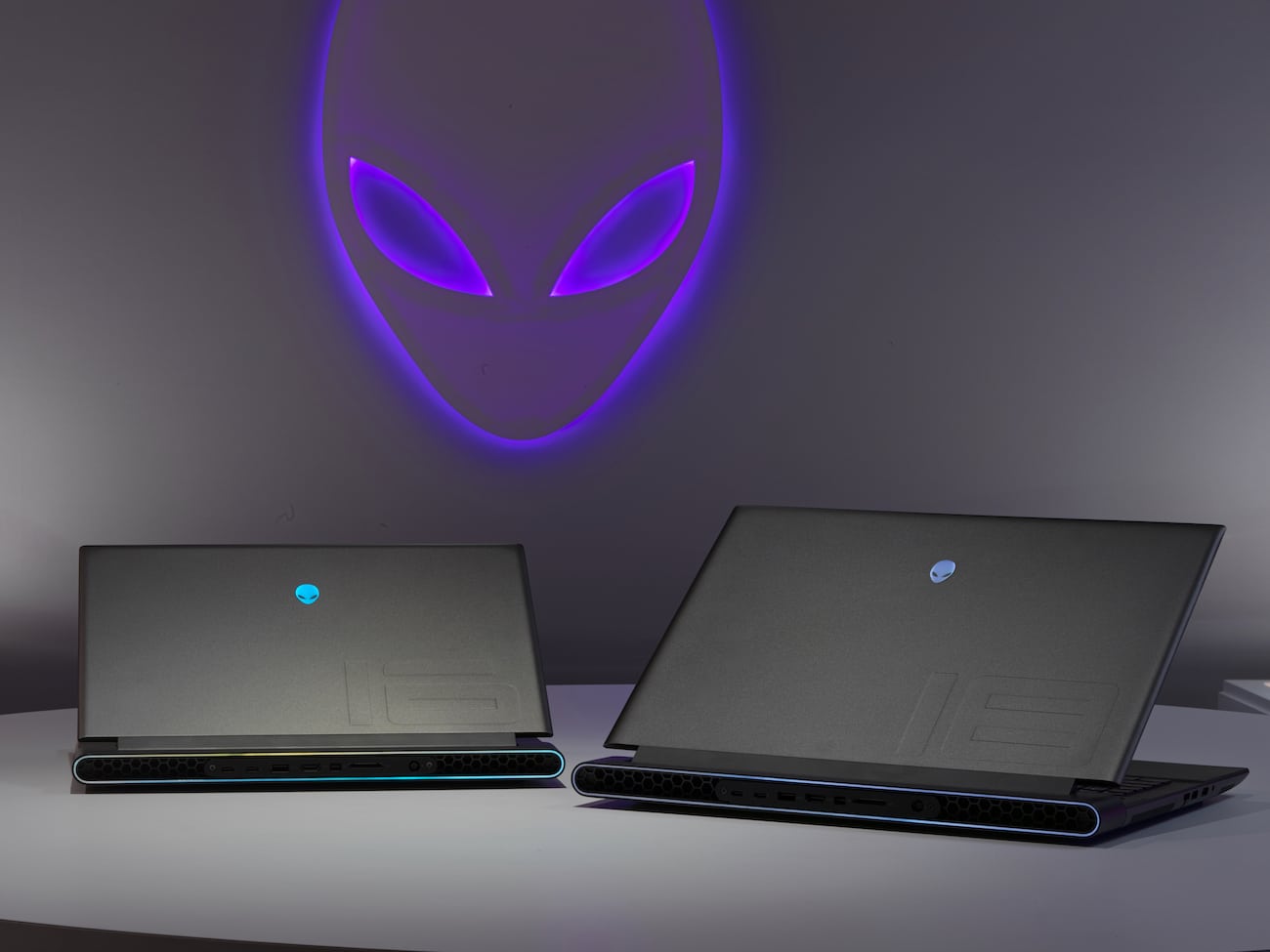 Dois notebooks gamer Alienware cinza com logo azul, lado a lado, em frente a um logo alienígena iluminado em roxo na parede (Alienware m18 e m16)