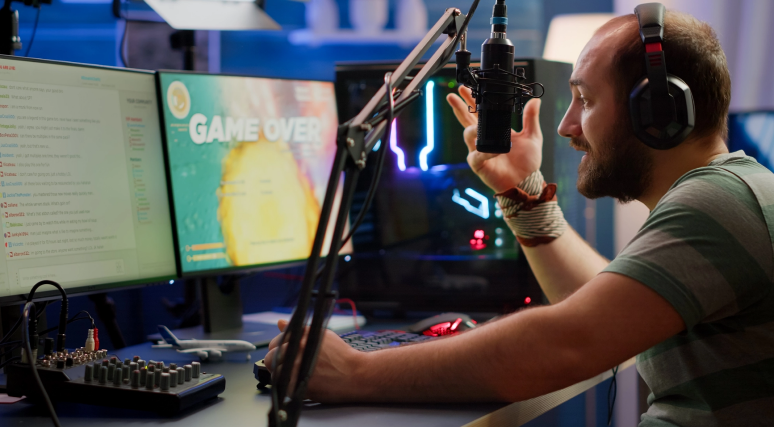 Streamer de barba e fones gamer falando ao microfone diante de monitores com tela "Game Over" e PC com iluminação RGB azul e roxa