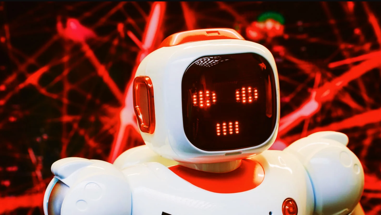 Robô branco e vermelho de brinquedo com tela de LEDs mostrando olhos e boca, diante de um fundo com linhas vermelhas brilhantes em forma de rede