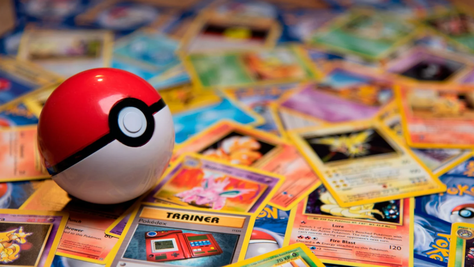 Pokébola vermelha e branca sobre um monte de cartas colecionáveis de Pokémon, com a carta Trainer Pokédex em destaque em primeiro plano