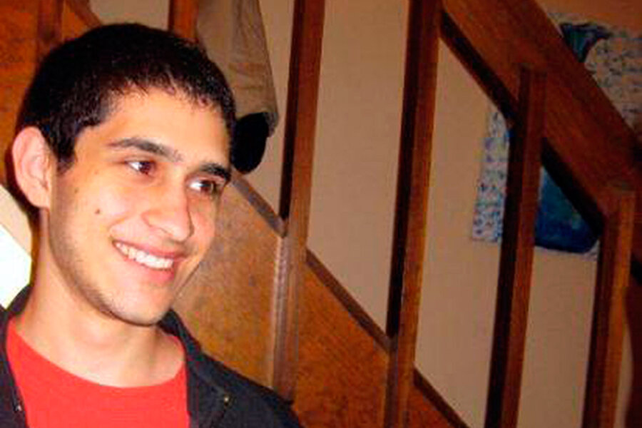 Jovem sorridente de camiseta vermelha e jaqueta escura, apoiado em uma escada de madeira com corrimão em uma casa