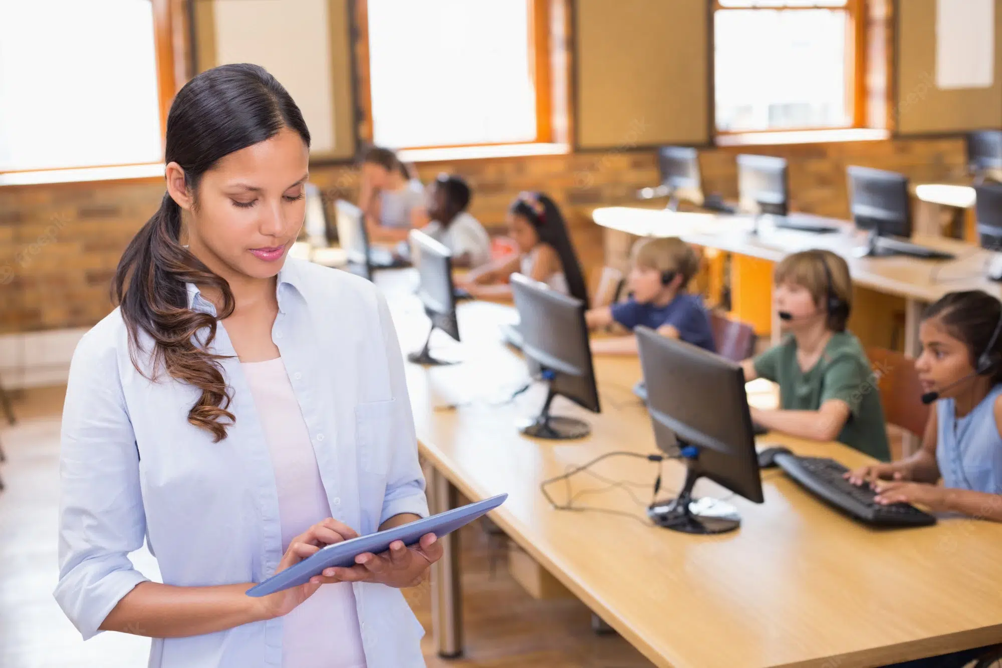 Professora com tablet observa alunos usando computadores com headsets em sala de informática