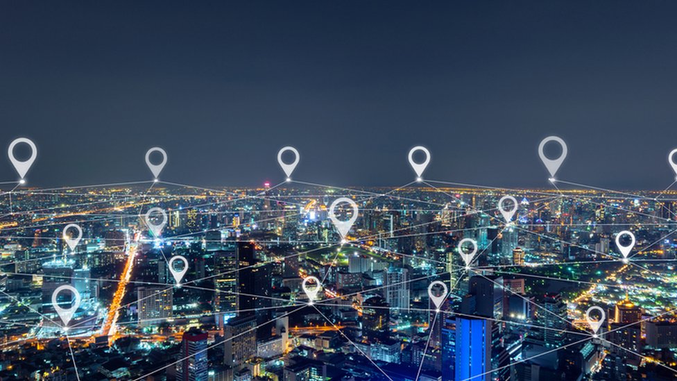 Vista noturna de uma cidade iluminada com ícones de localização e linhas conectando pontos sobre os prédios