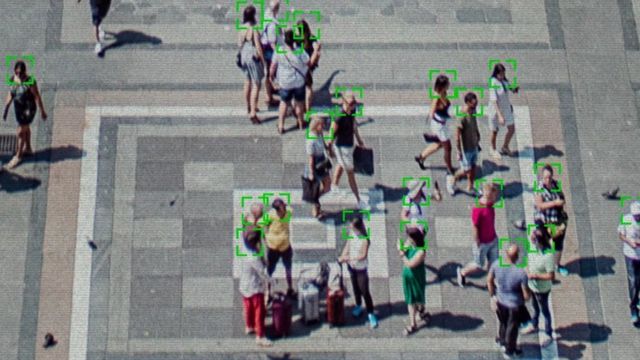 Pessoas caminhando em praça pública, com quadros verdes de reconhecimento facial sobre os rostos, ilustrando sistemas de identificação por câmeras