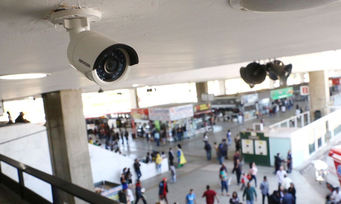 Câmera de segurança Hikvision instalada no teto sobre área de circulação movimentada, com pessoas caminhando em um terminal com lojas ao fundo
