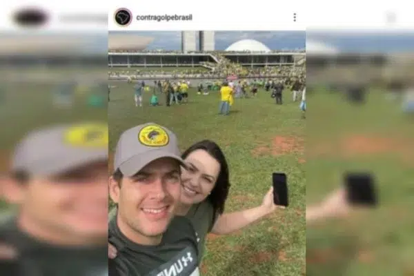 Casal sorrindo em selfie com o Congresso Nacional ao fundo e manifestantes no gramado, um deles com boné e camiseta de manga curta