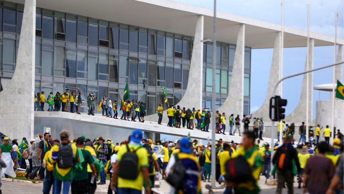 Multidão com roupas verdes e amarelas invade a rampa de um prédio de arquitetura modernista com colunas de concreto em Brasília, com bandeiras do Brasil ao alto